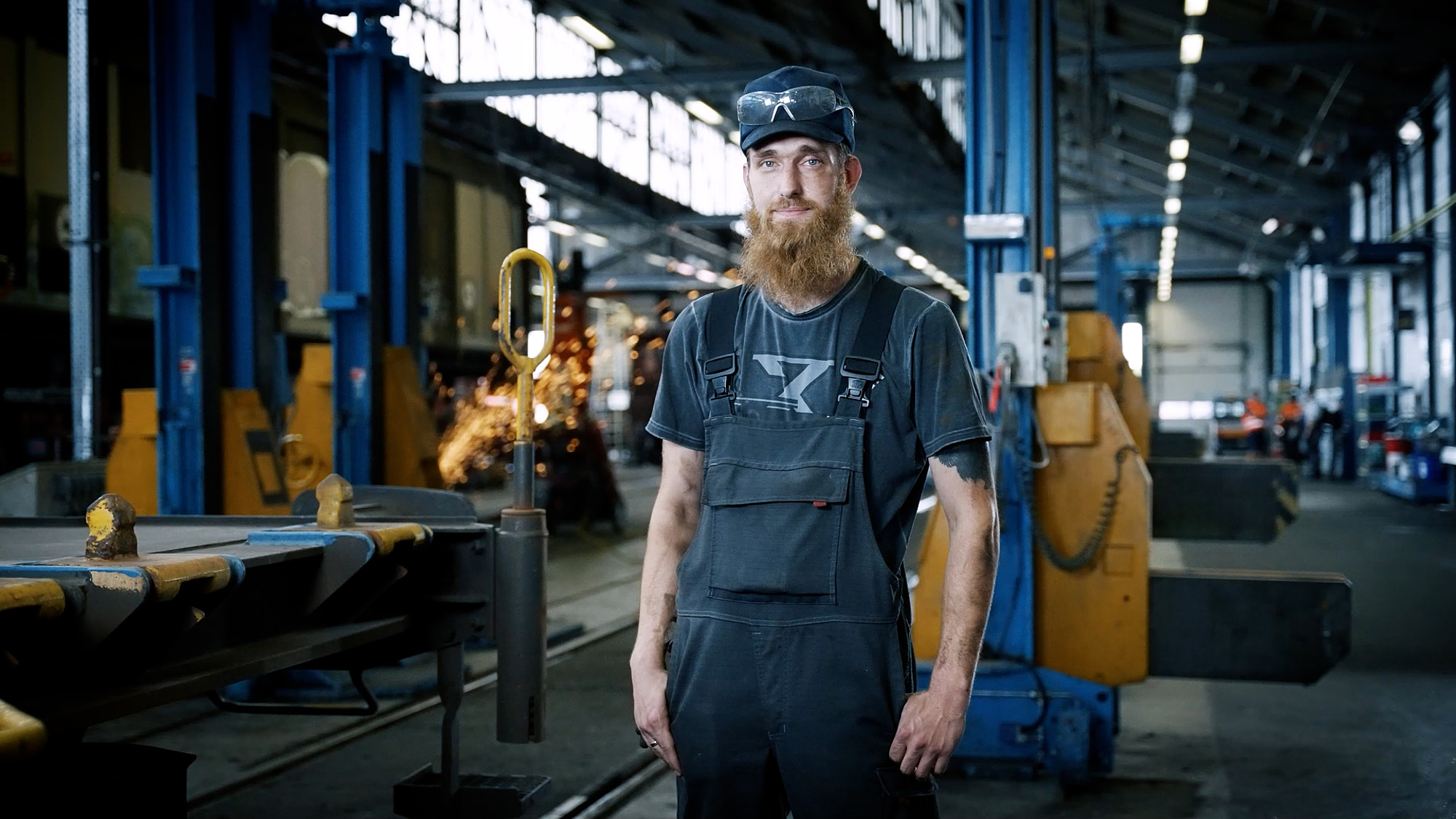 Handwerker mit Bart und Schutzbrille steht in Werkshalle zwischen Maschinen und Funkenflug.